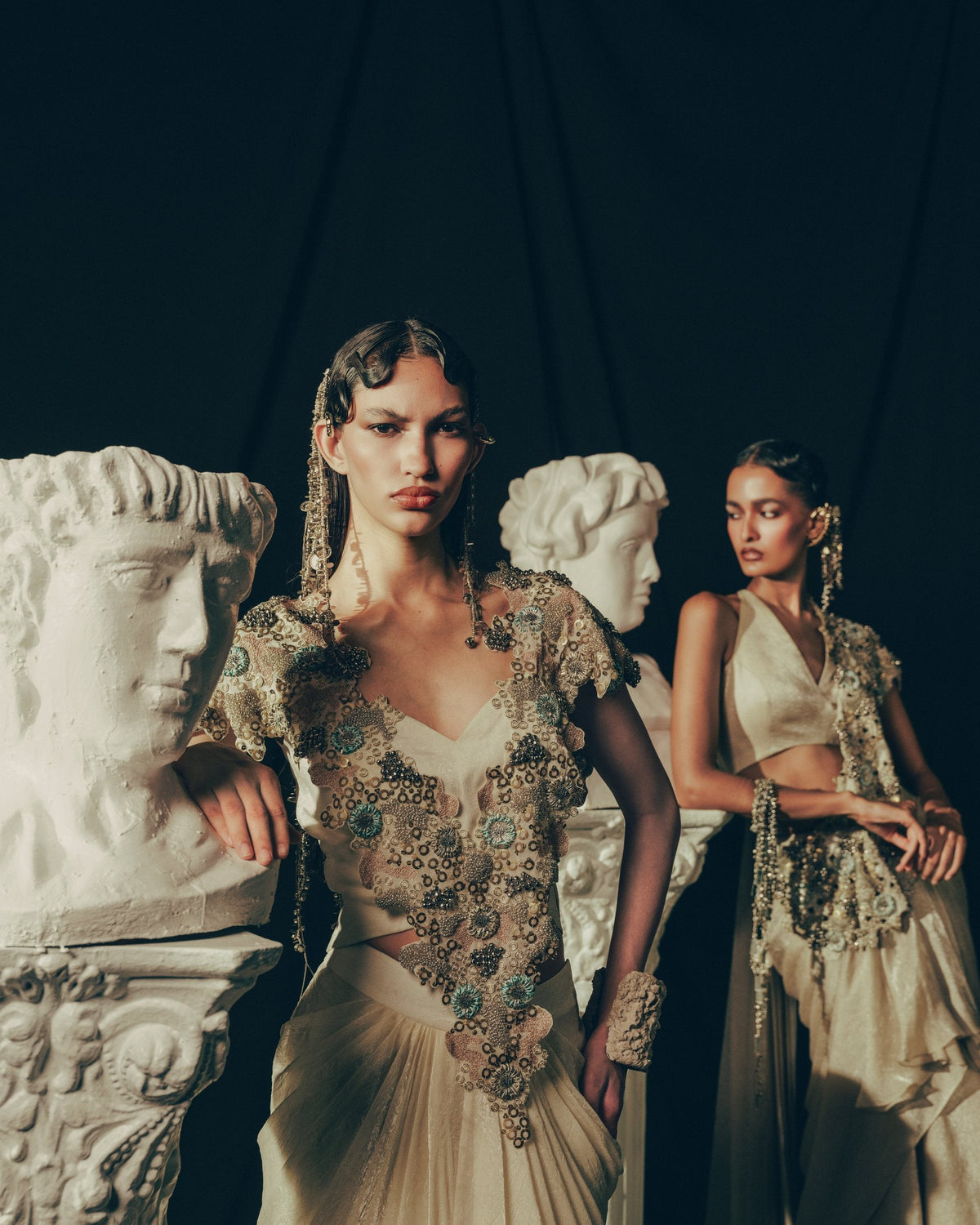Two models in ivory embroidered drapes beside plaster busts