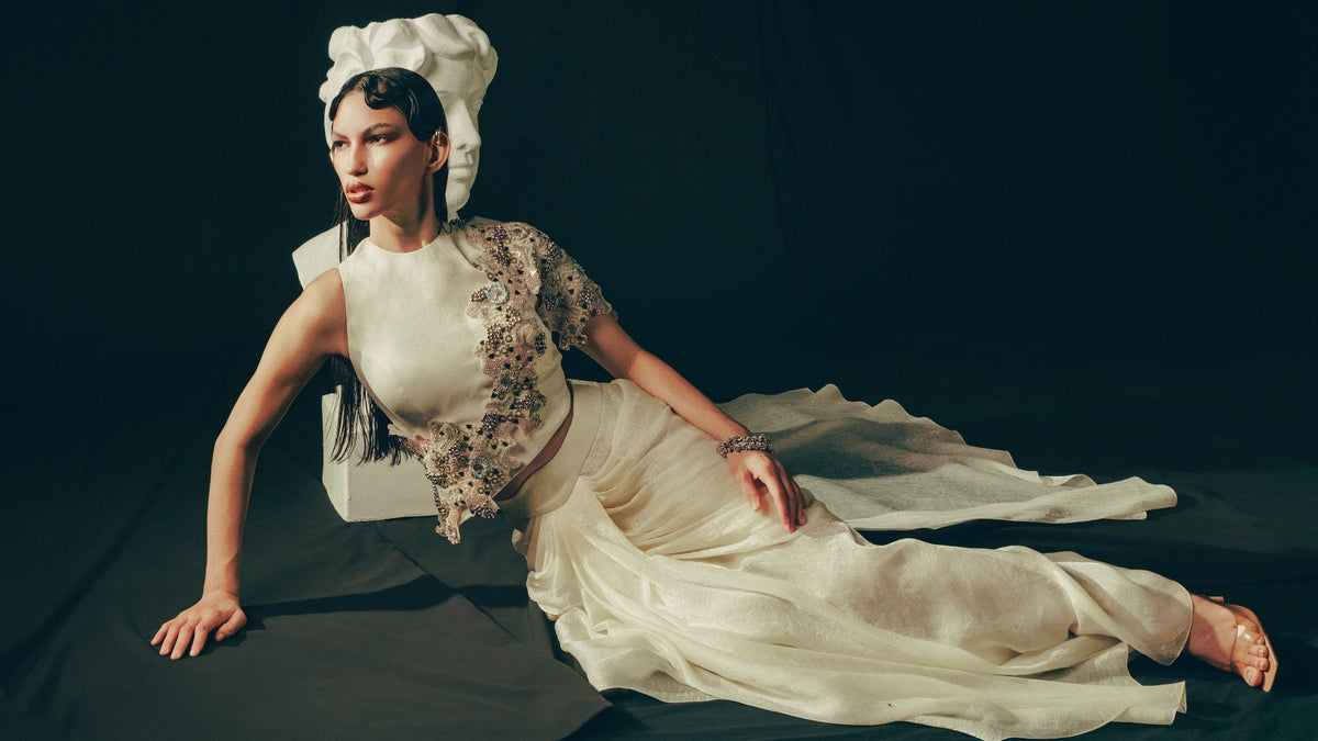 Model reclining in an ivory hand-embroidered drape set beside a plaster bust